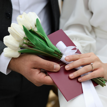 A close up of hands holding a bouquet of white tulips and a red booklet