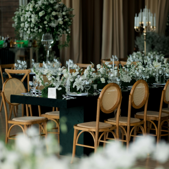 A table setting in a wedding venue showing brown wicker chairs, white flowers and candles on green tables.